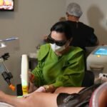 Profissional com óculos de proteção aplica laser transdérmico na perna de paciente deitada em clínica