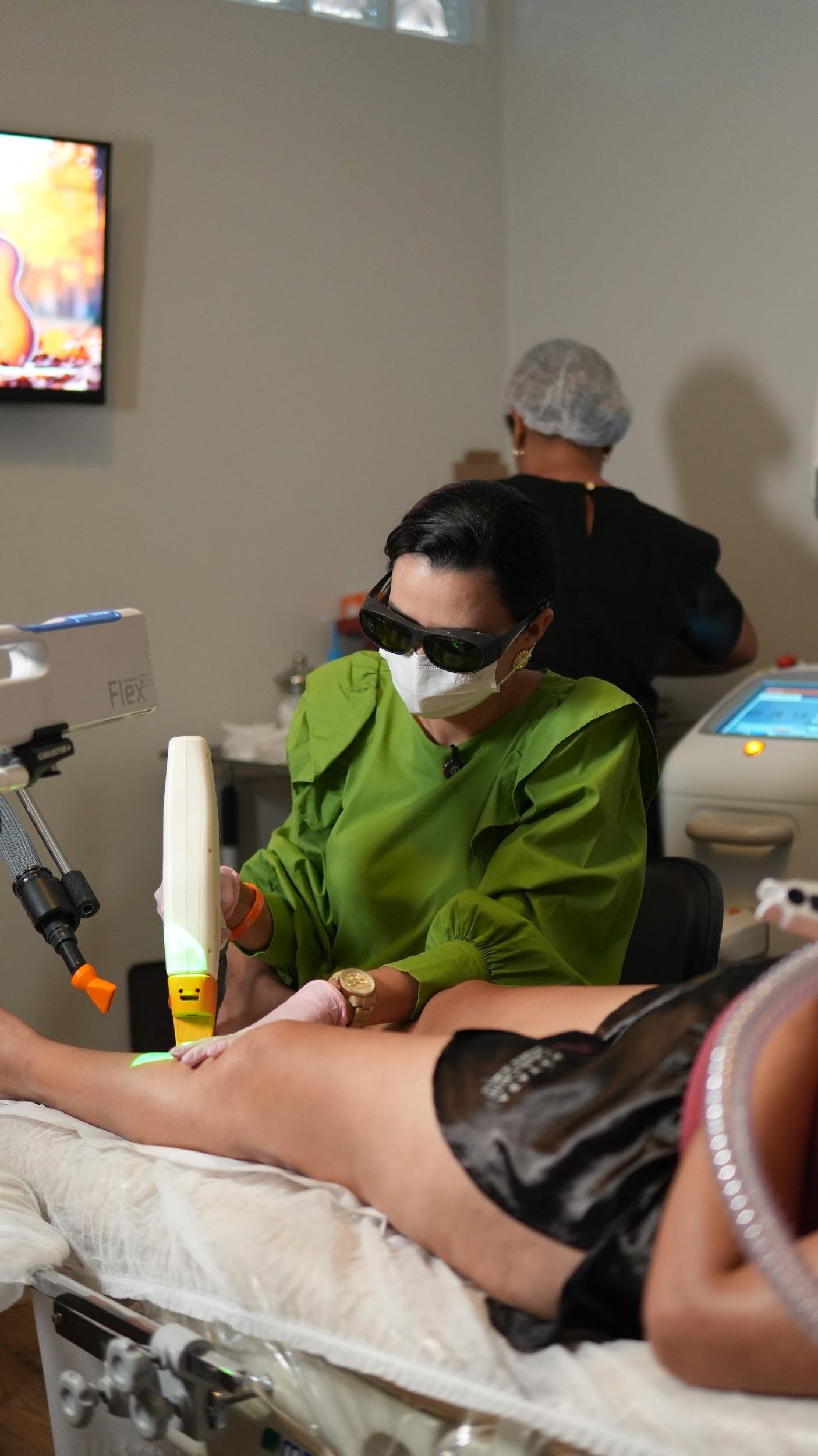 Profissional com óculos de proteção aplica laser transdérmico na perna de paciente deitada em clínica